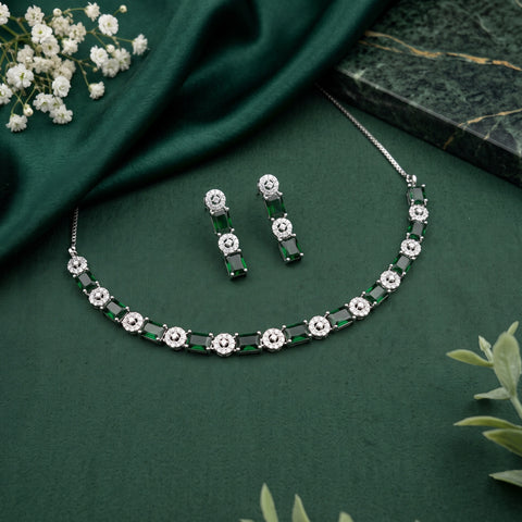 A silver-tone jewelry set featuring a necklace and matching drop earrings with alternating emerald-cut green stones and round clear stones, arranged on a dark green fabric backdrop with decorative flowers and a marble slab.