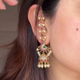 A close-up of a person's ear wearing a large gold-toned peacock-shaped jhumka earring decorated with pink stones, white pearls, and a small green accent, against dark brown hair.