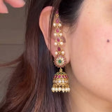 A close-up of a woman's ear wearing a long, ornate gold-toned dangle earring decorated with red stones, white pearls, and a green center stone.