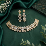 Gold-toned jewelry set with a matching necklace and drop earrings, arranged on dark green satin fabric with small white flowers and green leaves as styling props.