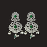 A close-up of a woman's ear wearing an ornate silver-toned traditional Indian earring with green emerald-cut stones and small dangling pearl-like beads, against a warm beige background.