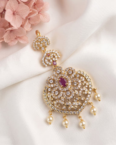 An ornate gold-toned traditional Indian bridal jewelry piece featuring intricate crystal pavé work, a central purple cabochon stone, and dangling white faux pearls, displayed on a draped white fabric with pink flowers in the corner.