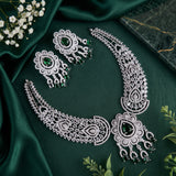 Silver-toned jewelry set with a necklace, earrings, and matching pendant arranged on dark green satin fabric, accented with small white flowers and green gemstone details.