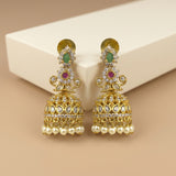 A pair of ornate gold-toned Indian jhumka earrings with green and red stones, clear pavé accents, and dangling pearl-like beads, displayed on a cream block against a beige background.