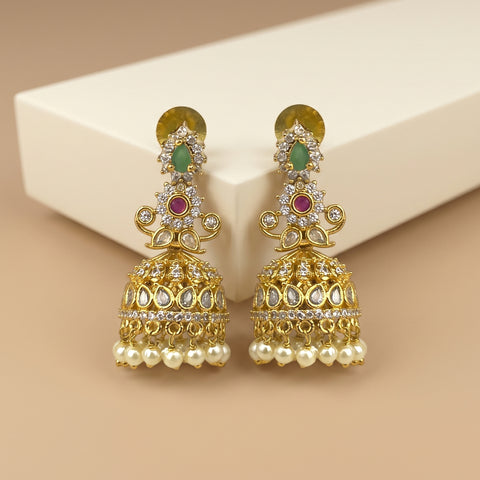 A pair of ornate gold-toned Indian jhumka earrings with green and red stones, clear pavé accents, and dangling pearl-like beads, displayed on a cream block against a beige background.