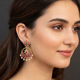 A close-up of a woman wearing a large gold-toned traditional Indian jhumka earring decorated with pink, green, and white stones and dangling pearl-like beads, against a dark gray background.