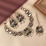 A traditional Indian jewelry set featuring a necklace, earrings, and a dangling charm, decorated with green enamel, pink stones, and clear rhinestones on a gold-toned base, arranged on a beige surface with a brown wooden frame in the corner.