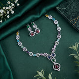 A luxury jewelry set featuring a V-shaped necklace and matching earrings with multi-colored gemstones in heart and teardrop shapes, displayed on a dark green satin fabric background with decorative floral elements.