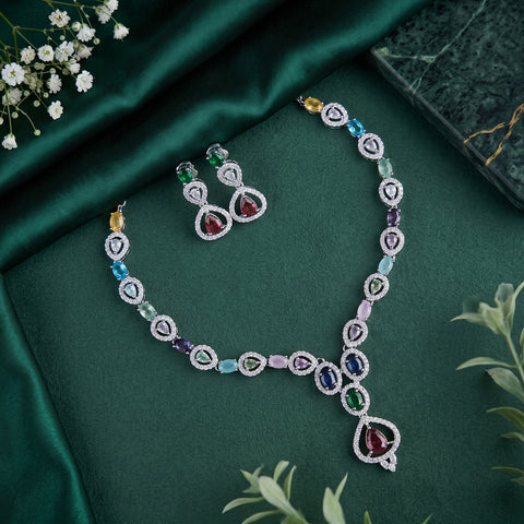 A luxury jewelry set featuring a V-shaped necklace and matching earrings with multi-colored gemstones in heart and teardrop shapes, displayed on a dark green satin fabric background with decorative floral elements.