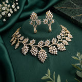 Gold-toned jewelry set with a leaf-shaped necklace, matching earrings, and a small matching box, arranged on dark green satin fabric with small white flowers and green leaves around it.