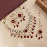 A gold-plated Indian jewelry set featuring a necklace, earrings, and a bracelet, embellished with clear crystals and deep red stones, displayed on a beige surface with a maroon box in the background.