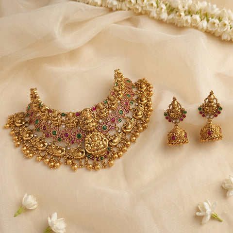 An ornate gold-toned traditional necklace with matching earrings, decorated with red and green stones, arranged on a soft cream fabric backdrop with white flowers.