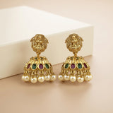 A pair of gold-toned Indian jhumka earrings with detailed filigree, green and purple stones, and dangling white faux pearls, displayed on a beige surface with a white block in the background.