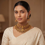 A woman wearing an ornate gold-toned traditional necklace with matching dangling earrings and a cream-colored saree, photographed against a softly blurred warm indoor background.