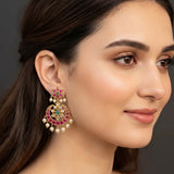 A close-up of a woman wearing a large, ornate gold-toned Indian-style chandelier earring decorated with green and pink stones and white pearl-like drops, against a dark gray background.