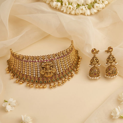 An ornate gold-toned traditional necklace with matching dangle earrings, decorated with rhinestones, small beads, and green and pink stone accents, arranged on a soft cream fabric backdrop with white flowers.
