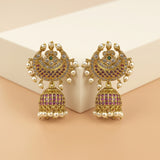A pair of ornate Indian jhumka earrings with gold-plated metal, purple stone inlay, and white faux pearls, displayed on a beige geometric block.
