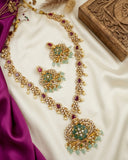 An ornate gold-toned jewelry set laid out on cream satin, including a long necklace, matching earrings, and a brooch, decorated with green stones, light blue bead drops, and purple gemstones. A carved wooden box and magenta fabric are visible as styling props.