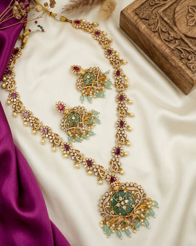 An ornate gold-toned jewelry set laid out on cream satin, including a long necklace, matching earrings, and a brooch, decorated with green stones, light blue bead drops, and purple gemstones. A carved wooden box and magenta fabric are visible as styling props.