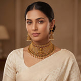 A woman wearing an ornate gold-toned traditional necklace with matching dangling earrings and a cream-colored saree, photographed in a softly lit indoor setting.