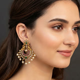 A close-up of a woman wearing a large, ornate gold-toned traditional Indian chandelier earring decorated with green and pink stones and white pearl-like drops, set against a dark gray background.