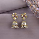 A pair of traditional Indian jhumka earrings with gold and silver floral tops, red center stones, and dangling pearl-like bells, displayed on a neutral mauve background with dried grass.