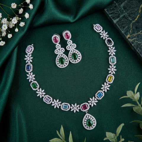 A silver-toned jewelry set featuring a necklace and matching drop earrings, decorated with multiple colorful gemstones including green, blue, pink, purple, and yellow, arranged on a dark green satin fabric background with small white flowers and green leaves.