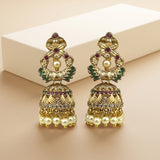 A pair of ornate gold-toned Indian jhumka earrings decorated with faux pearls, green stones, and small red gemstones, displayed on a beige surface with a cream-colored box in the background.