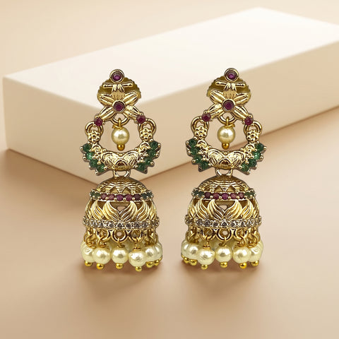 A pair of ornate gold-toned Indian jhumka earrings decorated with faux pearls, green stones, and small red gemstones, displayed on a beige surface with a cream-colored box in the background.