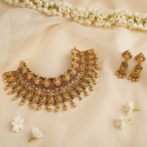 An ornate gold-toned traditional necklace with matching dangling earrings arranged on soft cream fabric, with small white flowers scattered around and a garland of white flowers along the top edge.