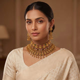 A woman wearing an ornate gold-toned traditional necklace with matching dangling earrings and a cream-colored saree, photographed in a softly lit indoor setting.
