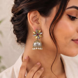 A close-up lifestyle shot of a woman wearing a traditional Indian jhumka earring. The earring features a gold and silver-toned flower-shaped stud with a small red stone and a dangling silver bell-shaped jhumka adorned with small white pearls.
