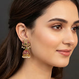 A close-up of a woman wearing a traditional Indian gold-plated jhumka earring with red and green enamel, white faux pearls, and a dangling bell-shaped charm.