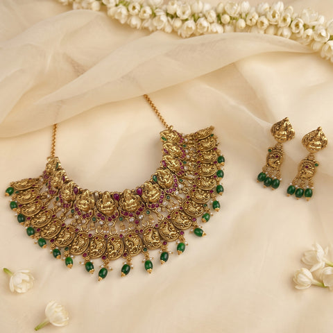 An ornate gold-toned traditional necklace with matching drop earrings, decorated with green beads, purple stones, and detailed motifs, arranged on a cream fabric backdrop with white flowers.