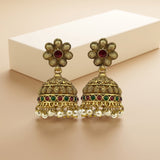 A pair of traditional Indian gold-plated jhumka earrings with red and green stone accents and white pearl fringe, displayed on a beige surface with a white box in the background.