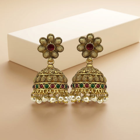 A pair of traditional Indian gold-plated jhumka earrings with red and green stone accents and white pearl fringe, displayed on a beige surface with a white box in the background.