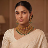A woman wearing an ornate gold-toned traditional necklace with green and red jewel accents, matching drop earrings, and a cream-colored saree, shown in a softly lit indoor setting.