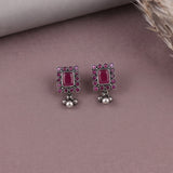 A pair of silver-toned rectangular dangle earrings with a central pink gemstone and a border of small pink stones, resting on a dusty mauve surface.