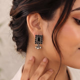 A close-up of a woman wearing a rectangular black and silver statement earring with dangling silver bead elements, shown on her ear and neck.
