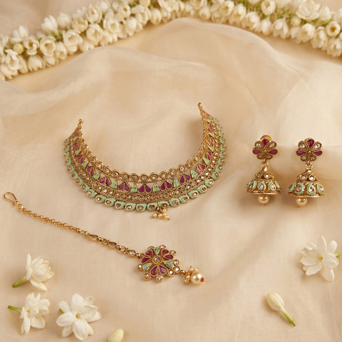 Gold-toned traditional Indian jewelry set including a choker necklace, matching earrings, and a bracelet, decorated with green and pink enamel details, arranged on a beige fabric backdrop with white flowers.