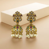 A pair of gold-plated Indian jhumka earrings with colorful enamel details and hanging pearl-like drops, displayed on a beige surface with a white rectangular block in the background.
