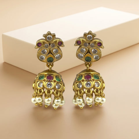 A pair of gold-plated Indian jhumka earrings with colorful enamel details and hanging pearl-like drops, displayed on a beige surface with a white rectangular block in the background.