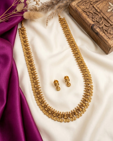 A gold-toned traditional necklace and matching earrings displayed on draped white fabric, with a purple satin cloth and a carved wooden box as styling props.