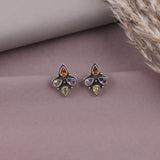 A pair of silver-toned stud earrings with four teardrop-shaped gemstones in amber, purple, yellow, and light green, displayed on a dusty mauve surface with dried grass in the corner.