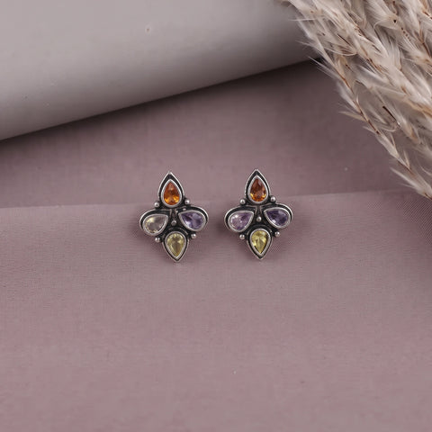 A pair of silver-toned stud earrings with four teardrop-shaped gemstones in amber, purple, yellow, and light green, displayed on a dusty mauve surface with dried grass in the corner.