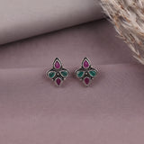 A pair of silver-toned stud earrings with teardrop-shaped green and pink enamel details, displayed on a dusty mauve surface with dried grass in the corner.