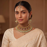A woman wearing an ornate gold-toned traditional necklace with green and pink stone accents, matching dangling earrings, and a maang tikka, paired with a light beige saree.