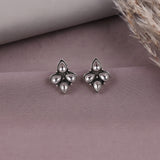 A pair of silver-toned floral stud earrings with teardrop-shaped clear stones, displayed on a textured mauve surface with a neutral cylindrical prop and dried grass in the background.