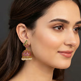 A close-up of a woman's ear wearing a gold-toned traditional Indian jhumka earring embellished with pink stones, green stones, and small white pearl-like beads.