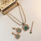 Gold-toned jewelry set with green stone accents, including a necklace, earrings, and a matching dangling piece, arranged on a light background beside an ornate decorative box.